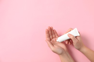 Photo of Woman applying cosmetic cream from tube onto her hand on pink background, top view. Space for text