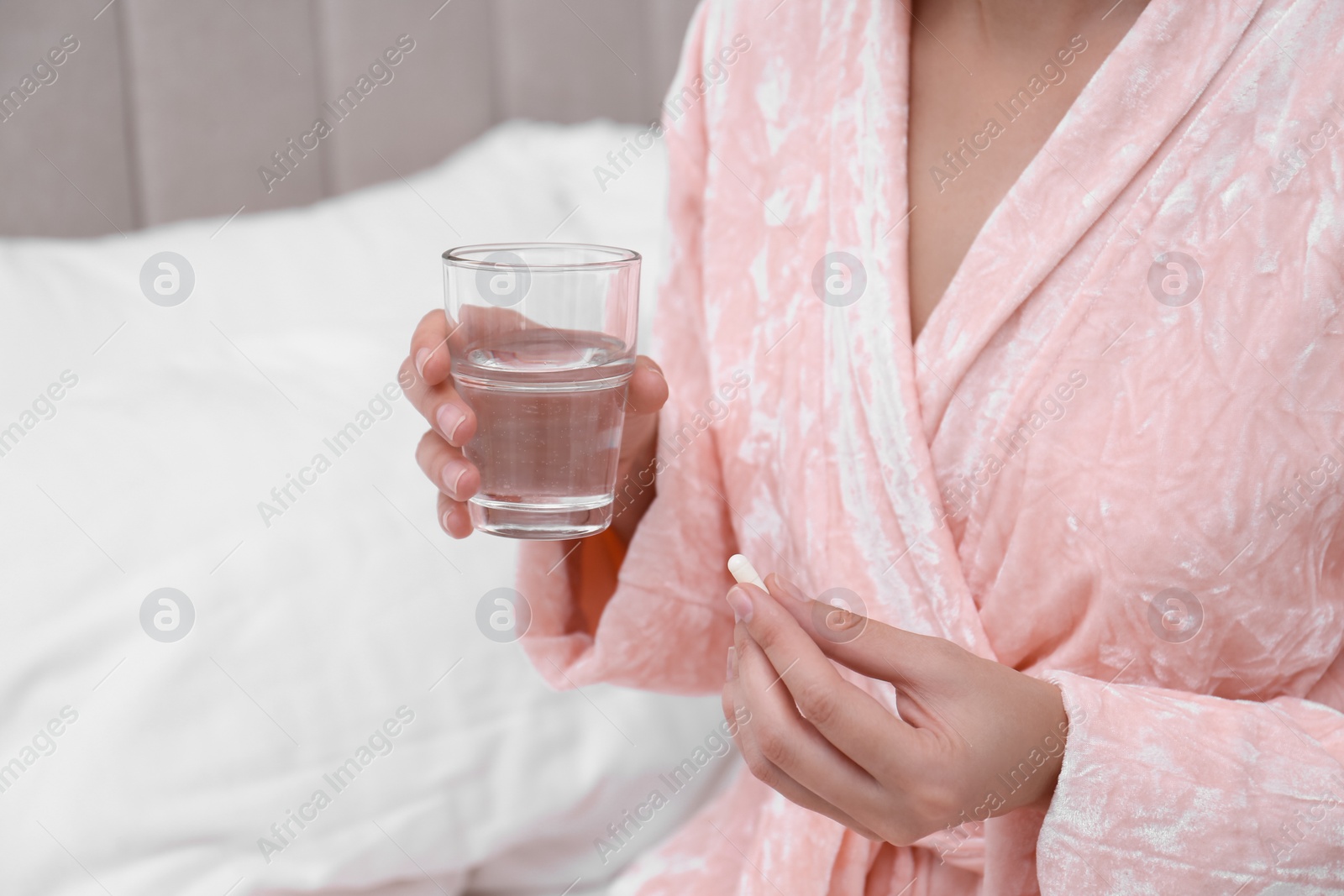 Photo of Young woman with glass of water taking pill in bedroom, closeup