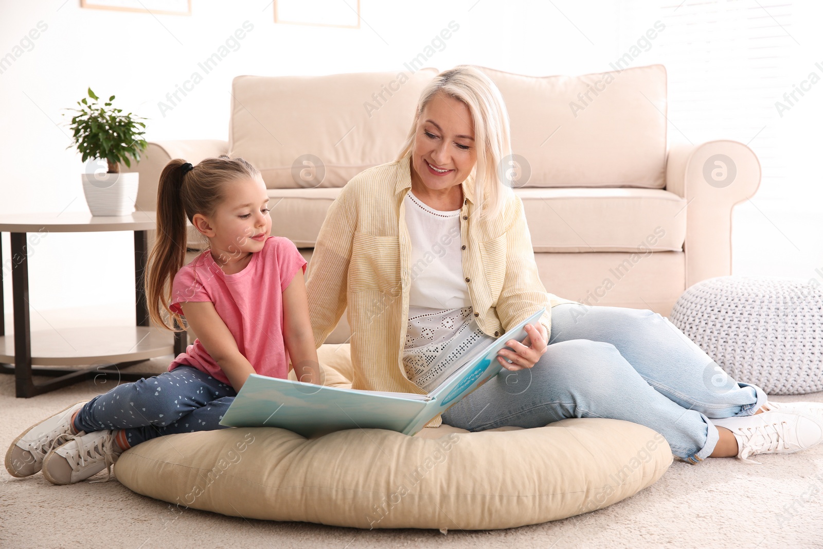 Photo of Mature woman with her little granddaughter reading book together at home