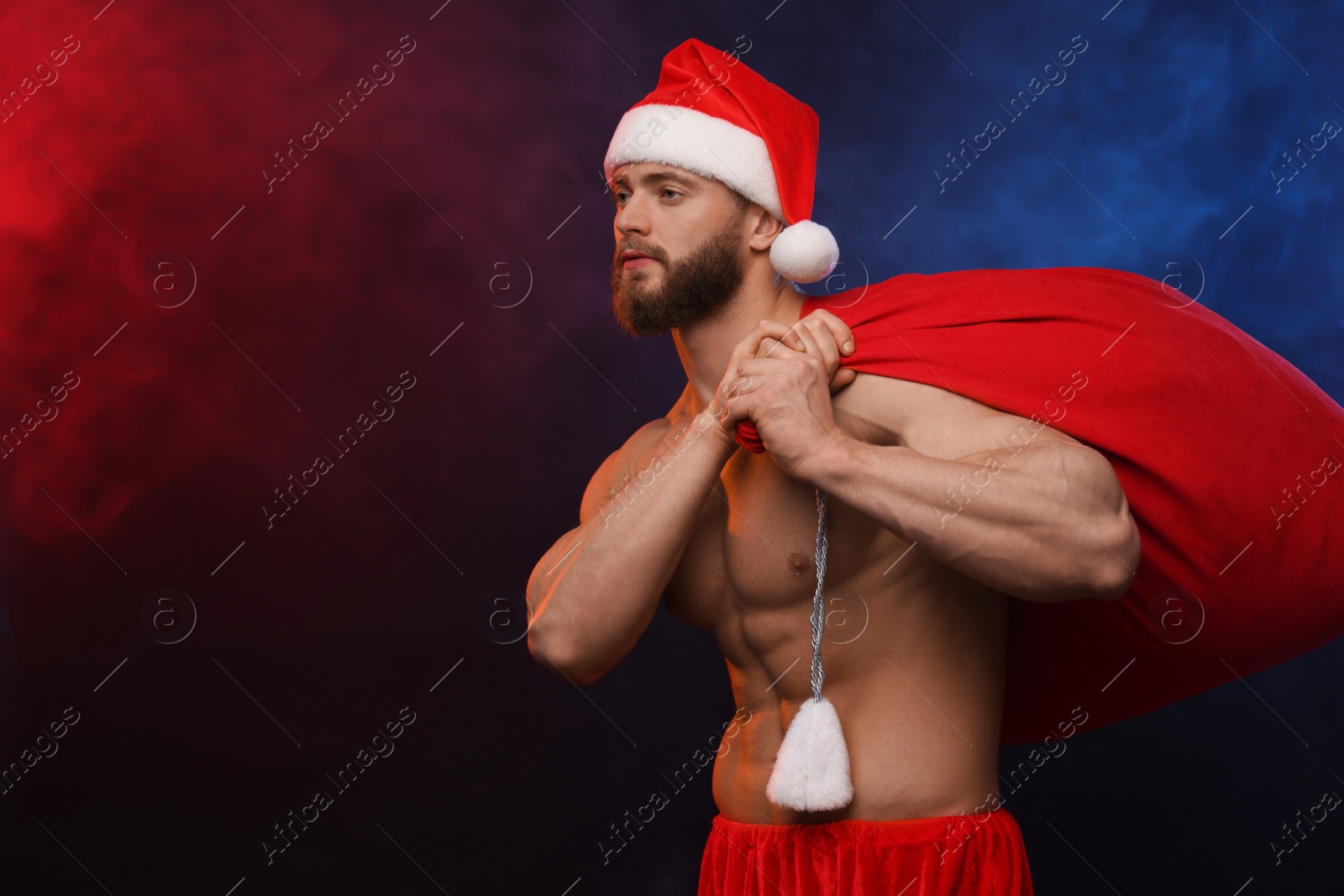Photo of Muscular young man in Santa hat holding bag with presents on color background, space for text