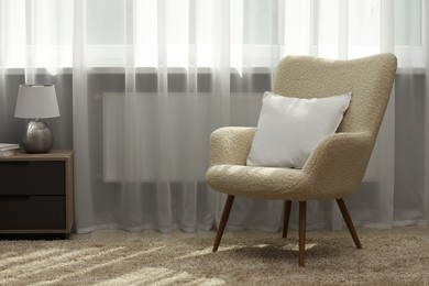 Soft white pillow on armchair and bedside table near window indoors