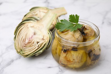 Photo of Jar of delicious artichokes pickled in olive oil and fresh vegetables on white marble table