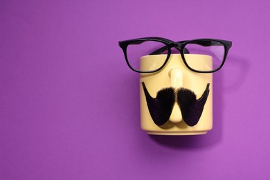 Photo of Man's face made of artificial mustache, cup and glasses on purple background, top view. Space for text