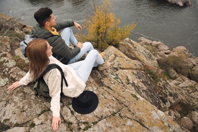 Couple of hikers with travel backpacks sitting on steep cliff near mountain river