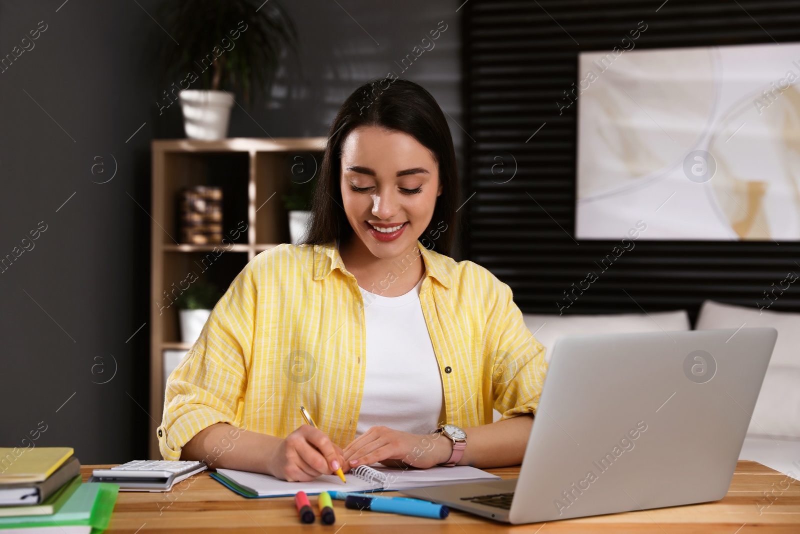 Photo of Young woman writing down notes during webinar at table in room