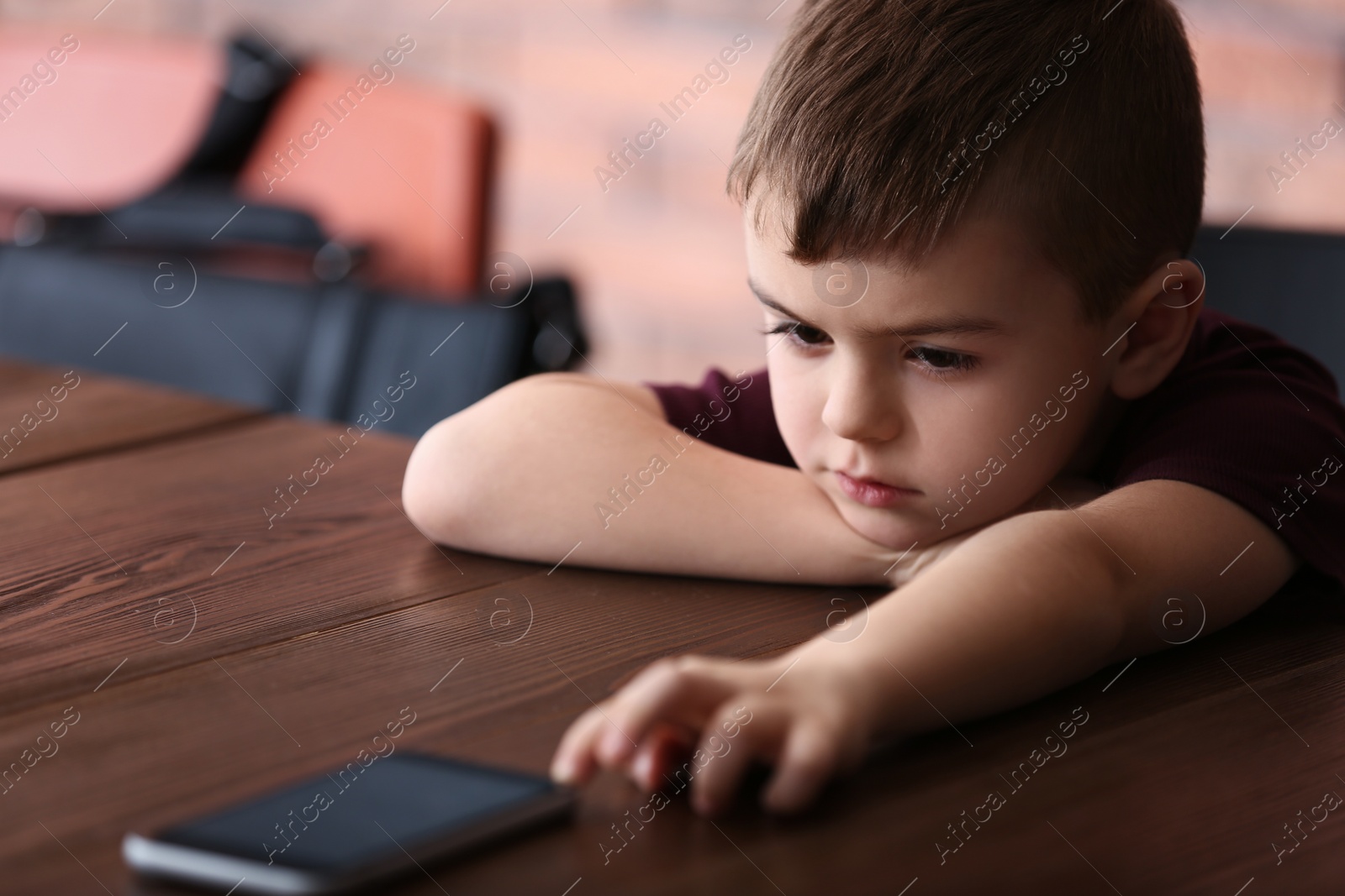 Photo of Sad little boy with mobile phone sitting at table indoors