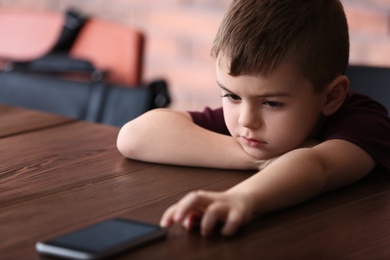 Sad little boy with mobile phone sitting at table indoors