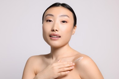 Beautiful young Asian woman applying body cream onto shoulder on light grey background