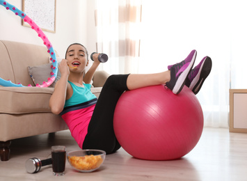 Photo of Lazy young woman with sport equipment eating junk food at home