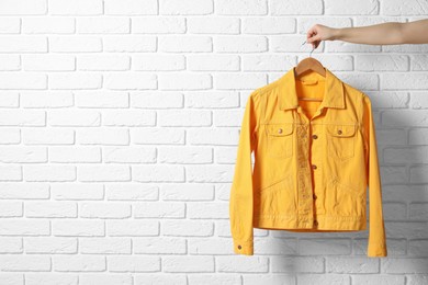 Photo of Woman holding hanger with yellow denim jacket near white brick wall, closeup. Space for text