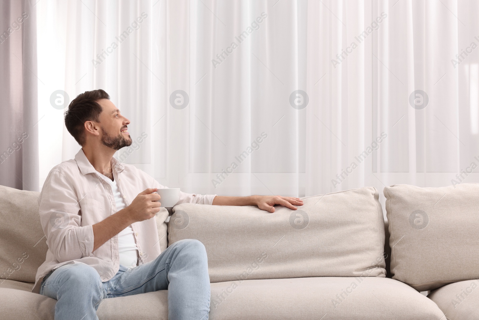 Photo of Happy man drinking coffee while resting on sofa near window with beautiful curtains in living room. Space for text