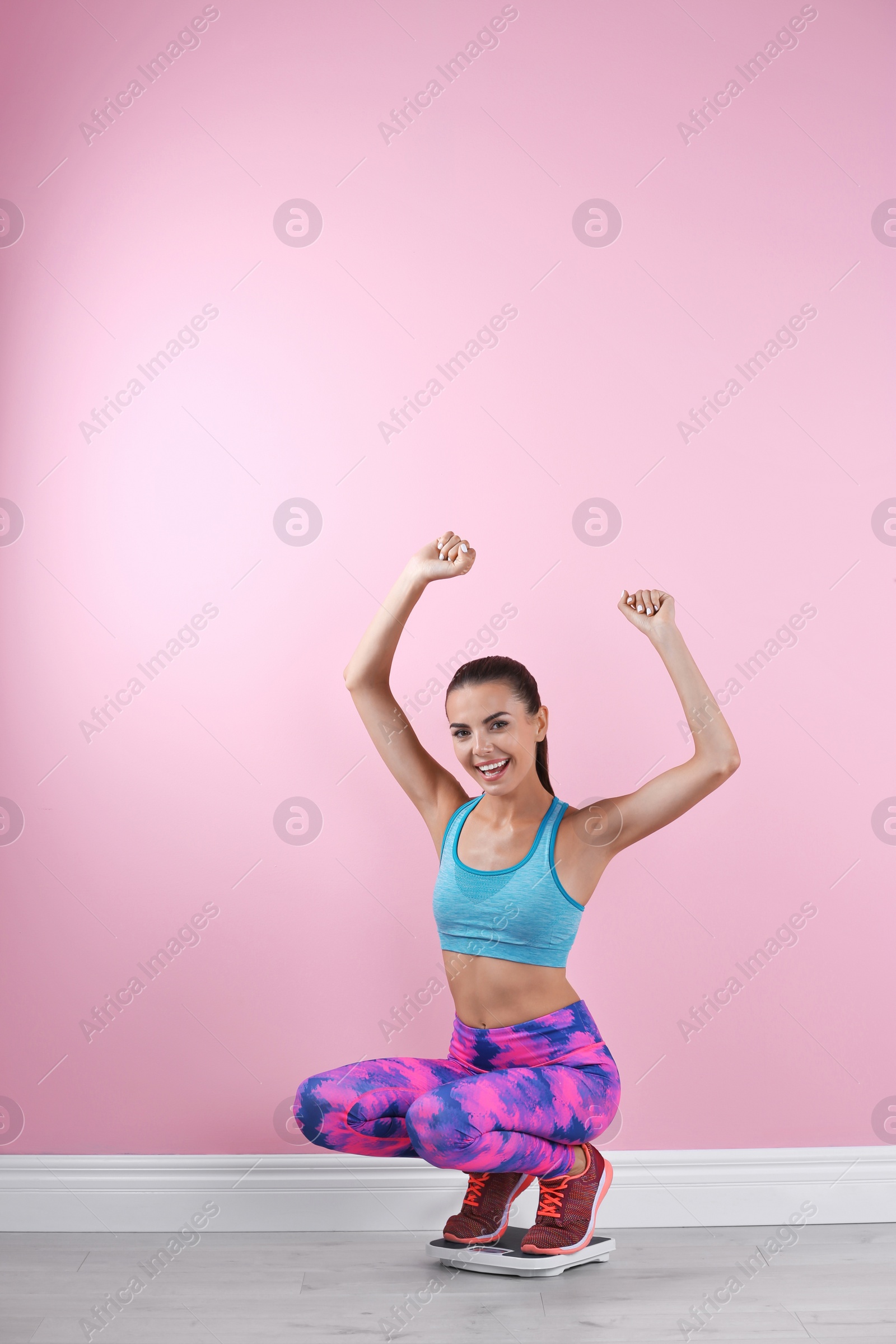 Photo of Happy young woman measuring her weight using scales near color wall. Weight loss motivation