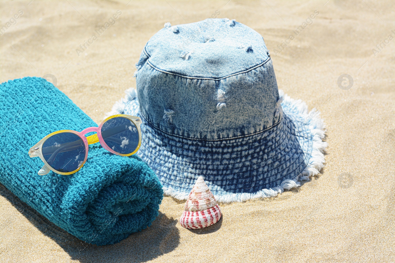 Photo of Towel with sunglasses, seashell and denim hat on sand outdoors. Beach accessories