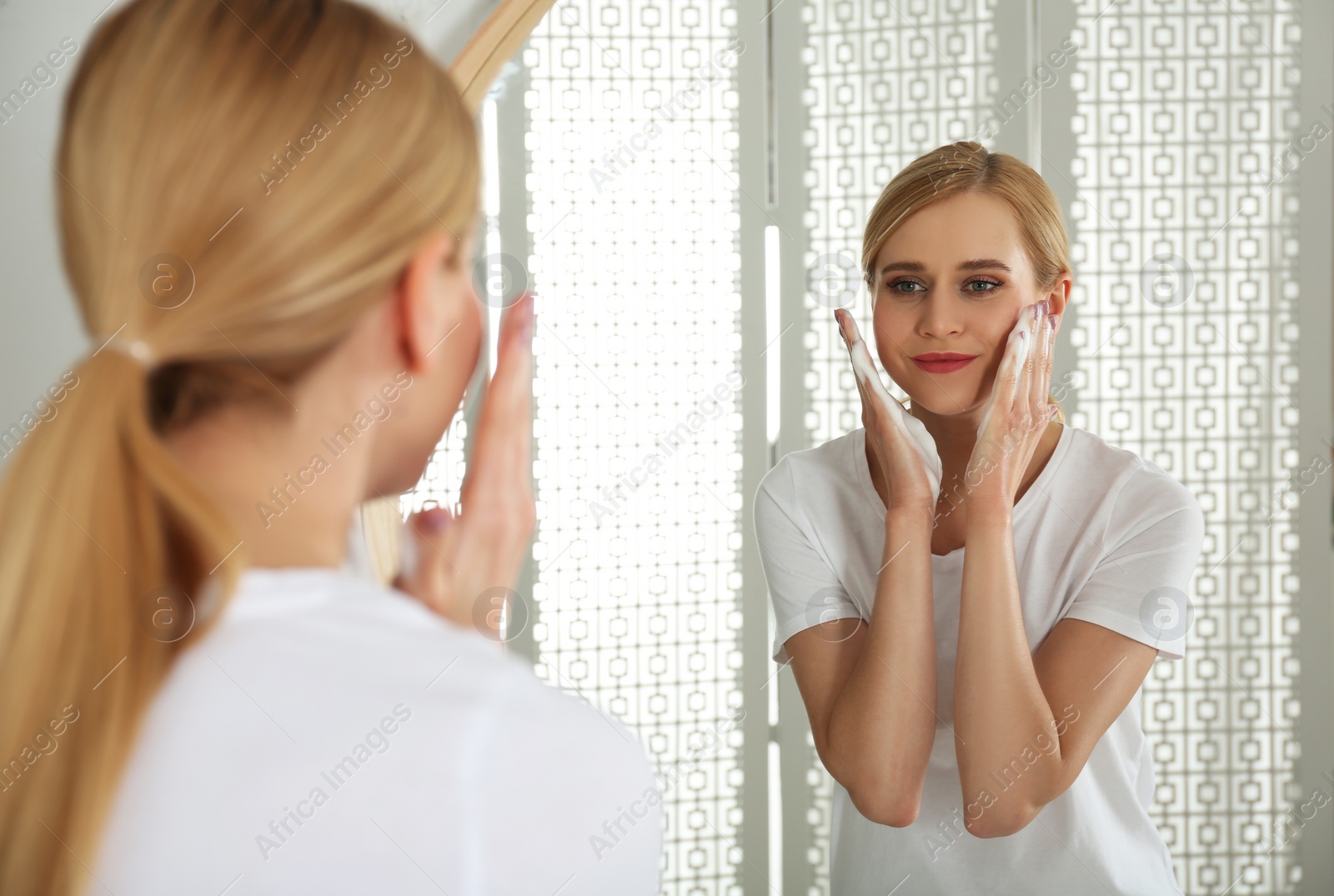 Photo of Happy young woman applying cleansing foam onto face near mirror in bathroom