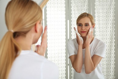 Happy young woman applying cleansing foam onto face near mirror in bathroom