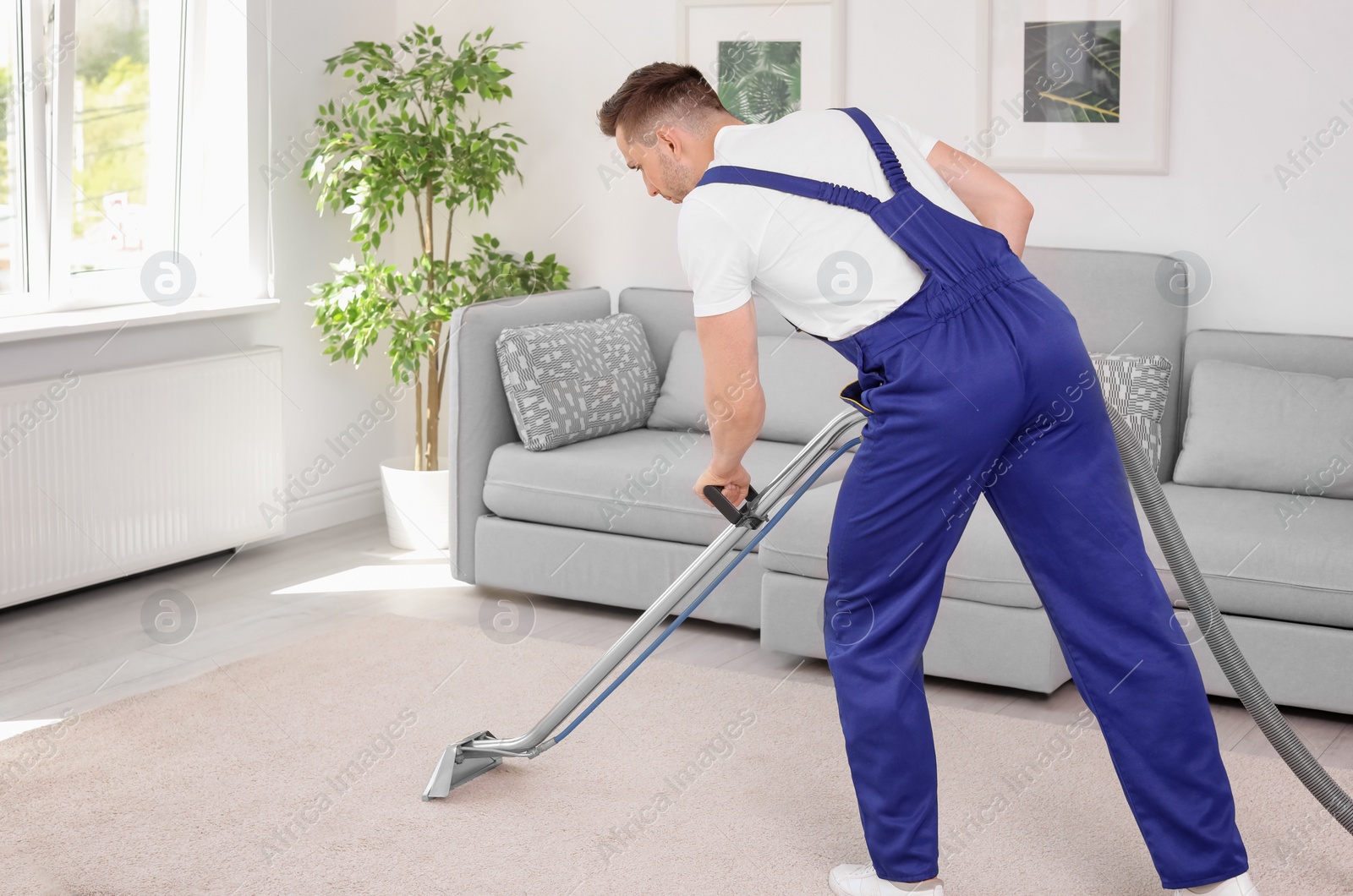 Photo of Male worker removing dirt from carpet with professional vacuum cleaner indoors