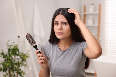 Photo of Emotional woman with brush examining her hair and scalp in bathroom. Dandruff problem