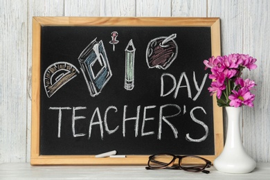 Photo of Chalkboard with inscription TEACHER'S DAY and vase of flowers on wooden table against white wall