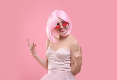 Pink look. Beautiful woman in wig and bright sunglasses showing rock sign on color background