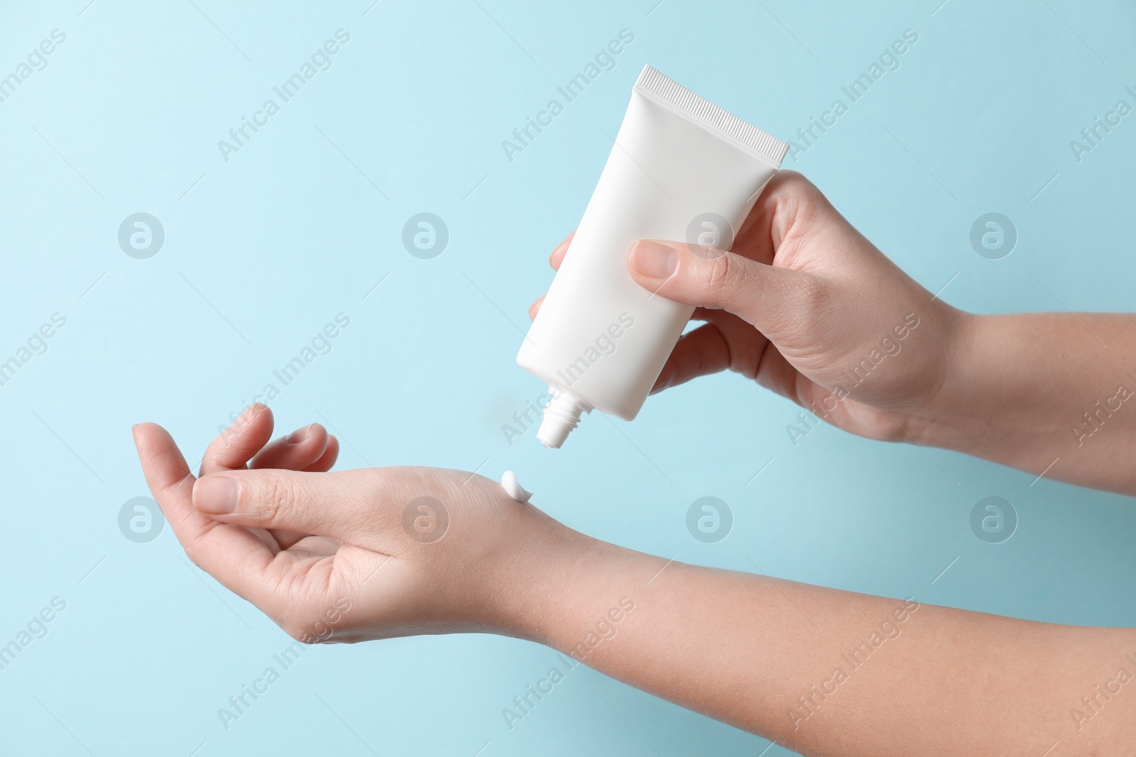 Photo of Woman applying cosmetic cream from tube onto her hand on light blue background, closeup