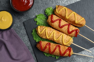 Photo of Delicious deep fried corn dogs with lettuce and sauces on grey table, flat lay