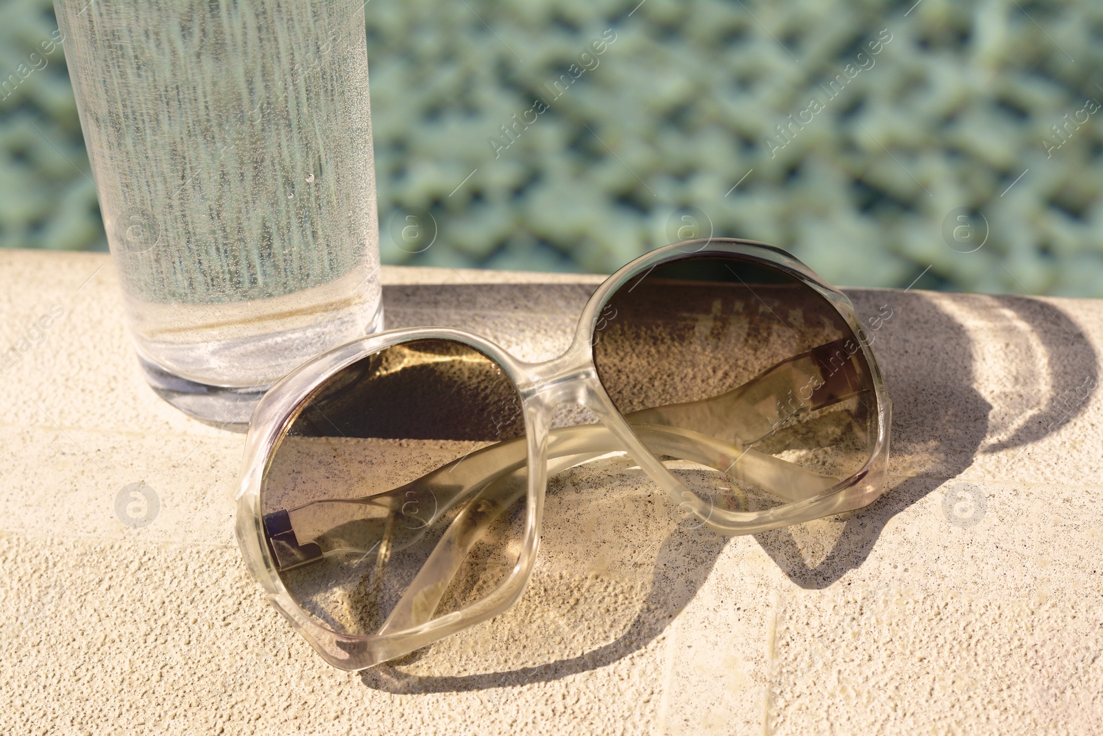 Photo of Stylish sunglasses and glass of water near outdoor swimming pool on sunny day, closeup