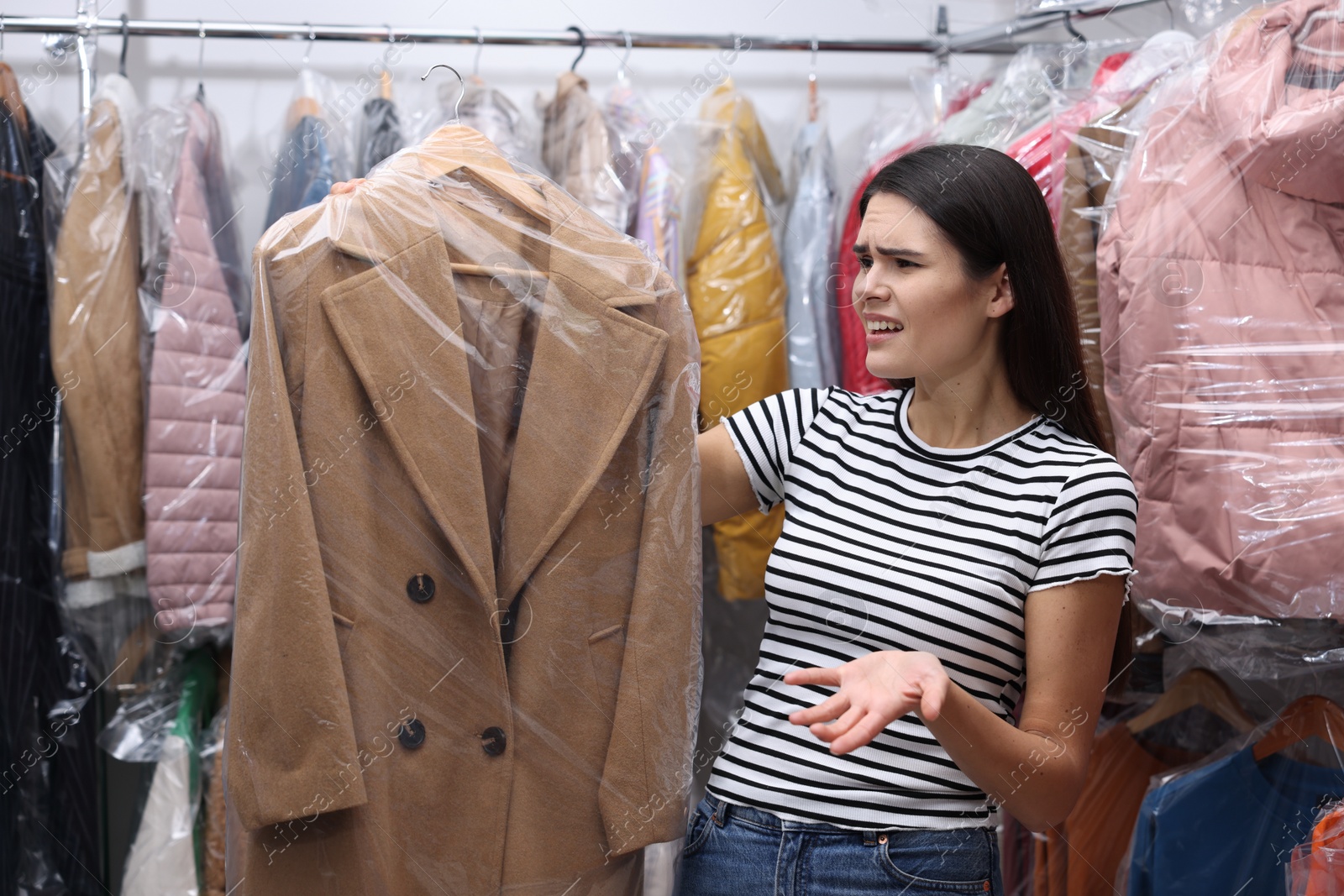 Photo of Dry-cleaning service. Displeased woman holding hanger with coat in plastic bag near racks with clothes indoors