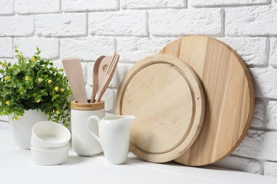 Photo of Cutting boards, holder with kitchen utensils, dishware and houseplant on white table near brick wall
