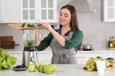 Beautiful young woman adding broccoli into blender with ingredients for smoothie at white table in kitchen