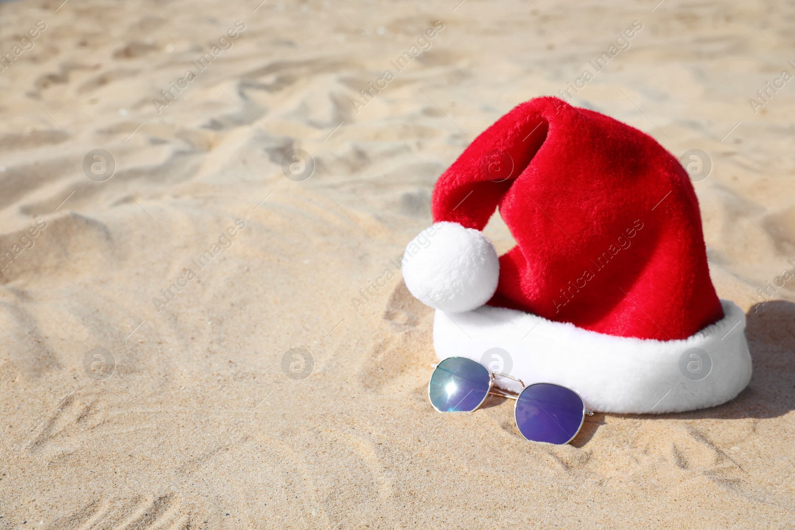 Photo of Santa's hat and sunglasses on sandy beach, space for text. Christmas vacation