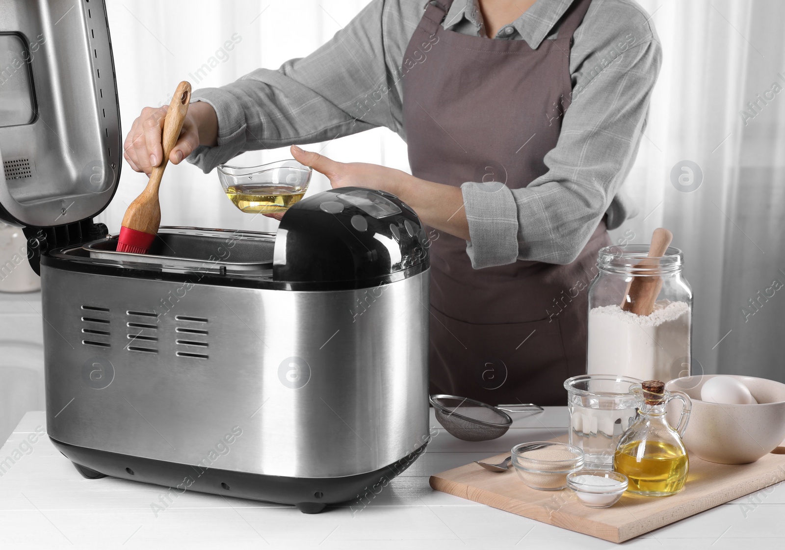 Photo of Woman applying oil onto breadmaker at white wooden table indoors, closeup