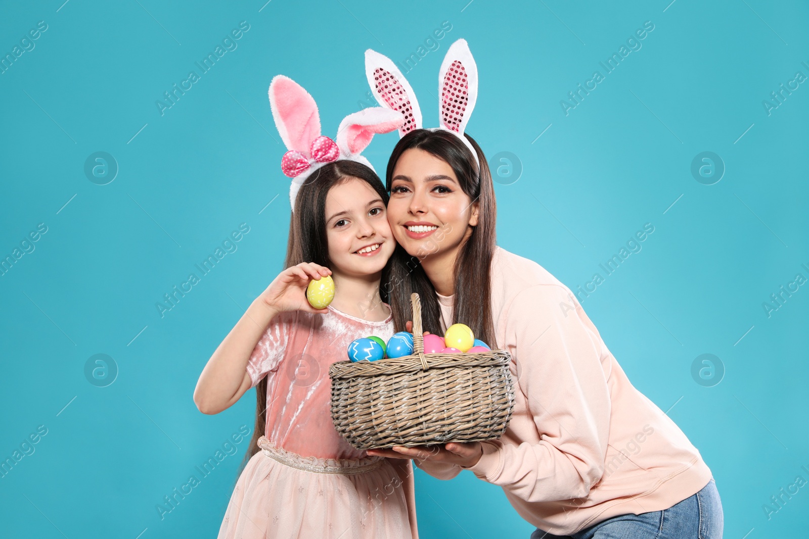 Photo of Mother and daughter in bunny ears headbands with basket of Easter eggs on color background