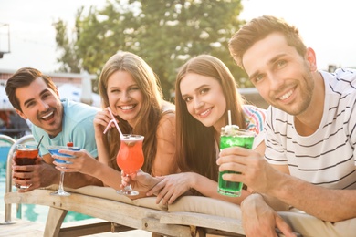 Happy young friends with fresh summer cocktails relaxing near swimming pool