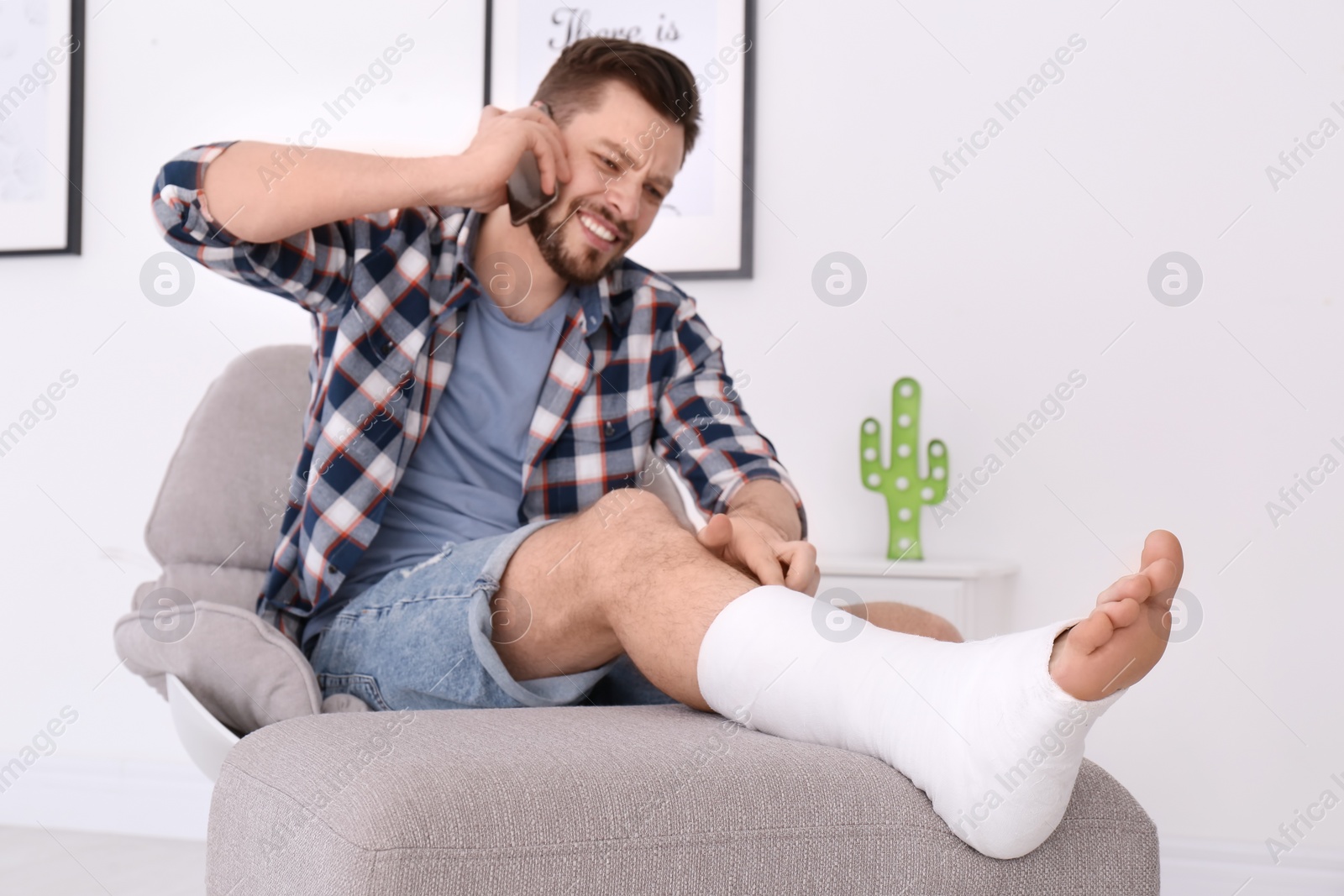 Photo of Man with broken leg in cast talking on mobile phone while sitting in armchair at home