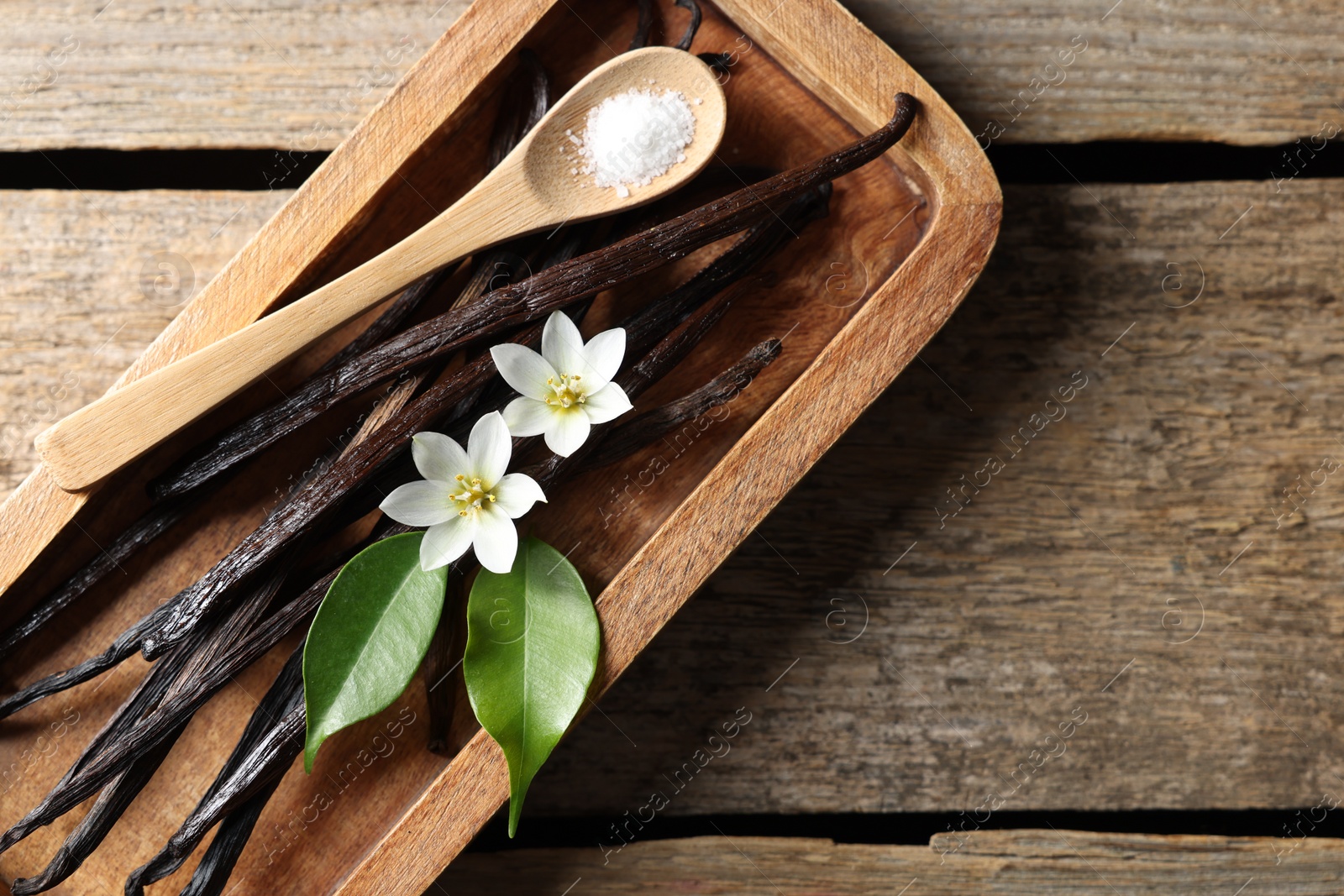 Photo of Vanilla pods, flowers, leaves and spoon with sugar on wooden table, top view