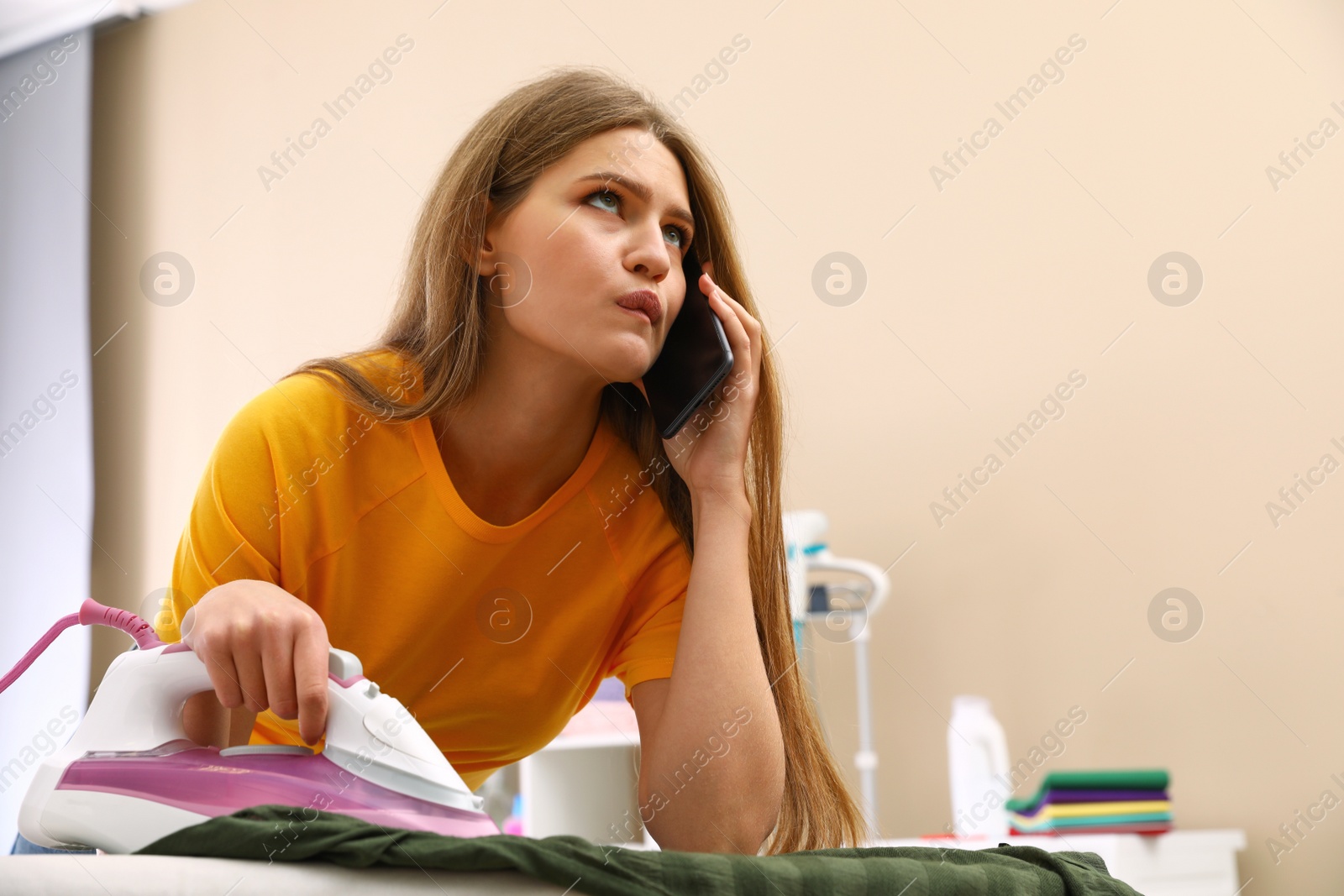 Photo of Thoughtful woman talking on phone while ironing clothes at home. Space for text