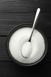 Granulated sugar in bowl and spoon on black wooden table, top view