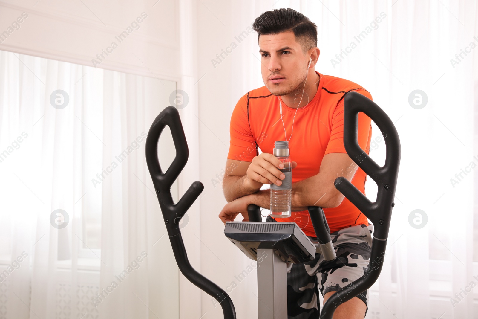Photo of Man with bottle resting after training on modern elliptical machine indoors