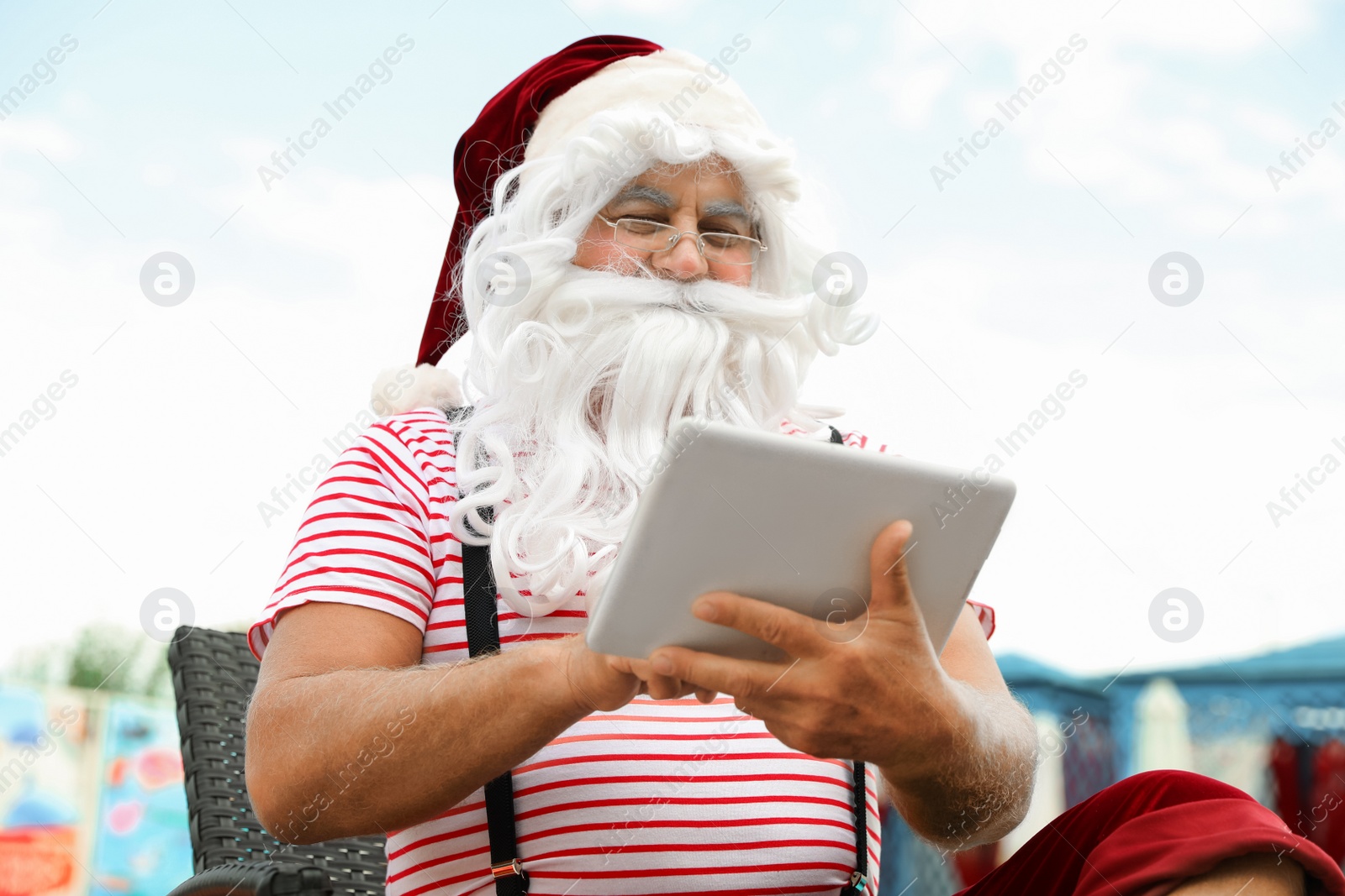 Photo of Authentic Santa Claus with tablet working on lounge chair at resort