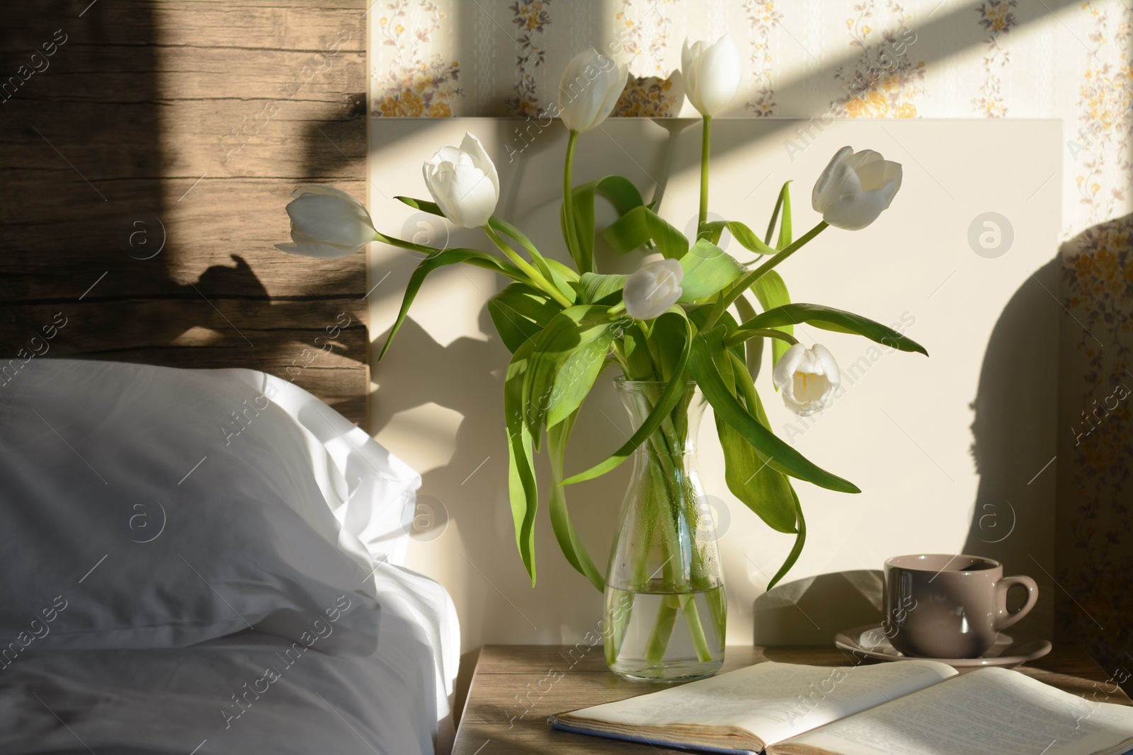 Photo of Beautiful white tulip bouquet, cup of drink and open book on nightstand in bedroom