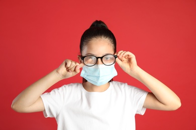 Little girl wiping foggy glasses caused by wearing medical face mask on red background. Protective measure during coronavirus pandemic