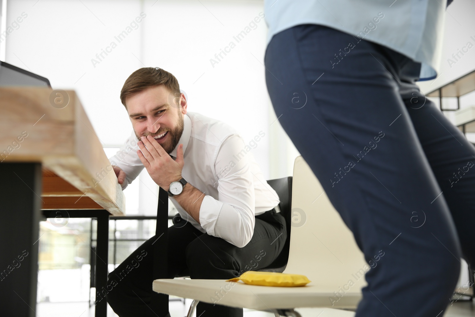 Photo of Young man putting whoopee cushion on chair while his colleague sitting down in office, closeup. Funny joke