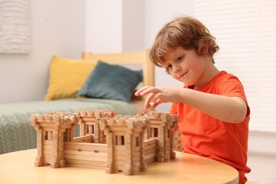 Cute little boy playing with wooden fortress at table in room. Child's toy