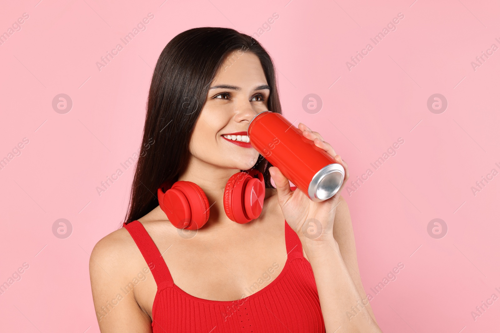 Photo of Beautiful young woman with headphones drinking from tin can on pink background