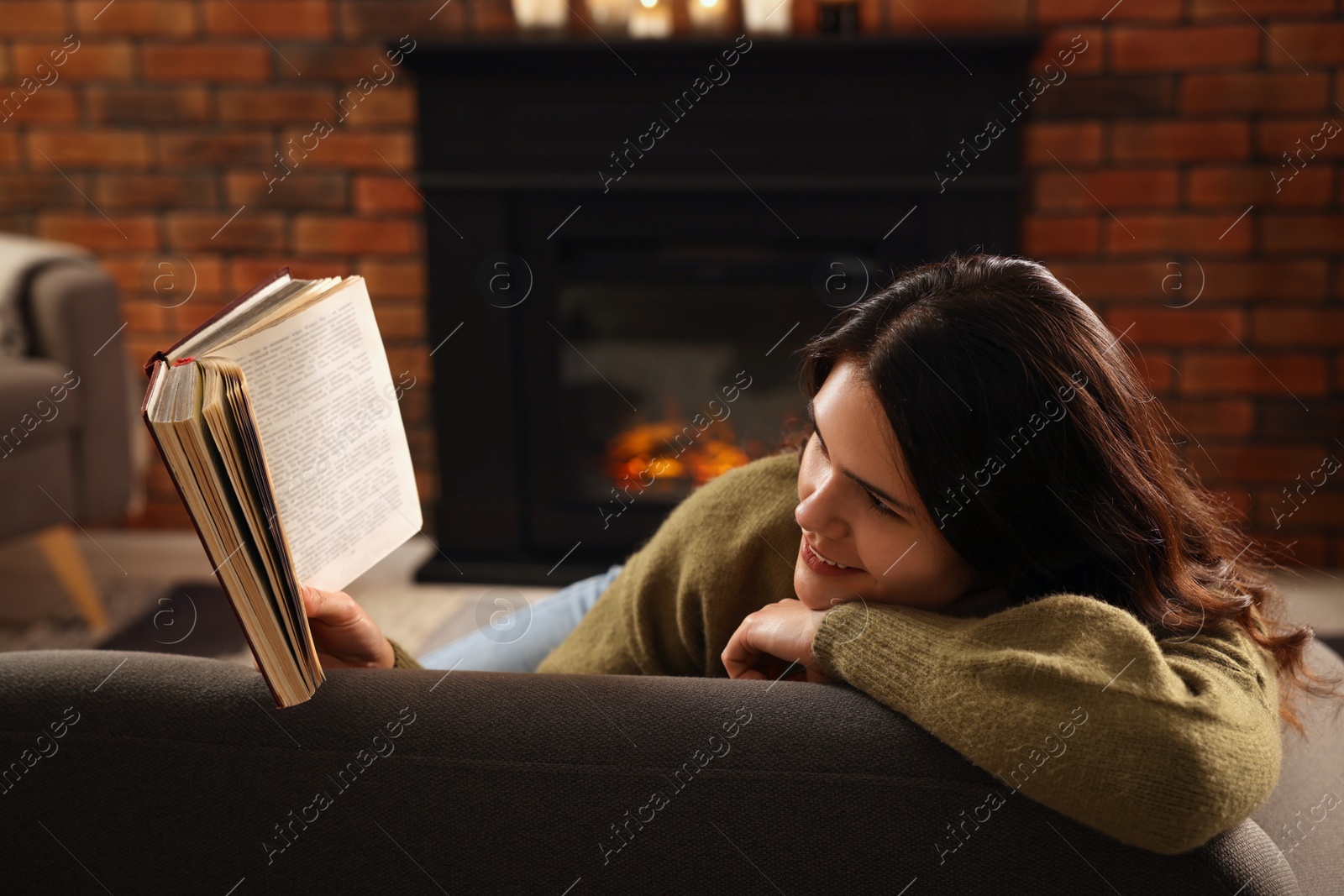 Photo of Young woman reading book on sofa near fireplace at home