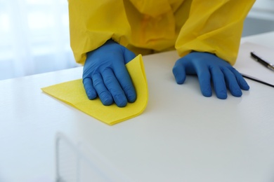 Photo of Employee in protective suit and gloves sanitizing table indoors, closeup. Medical disinfection