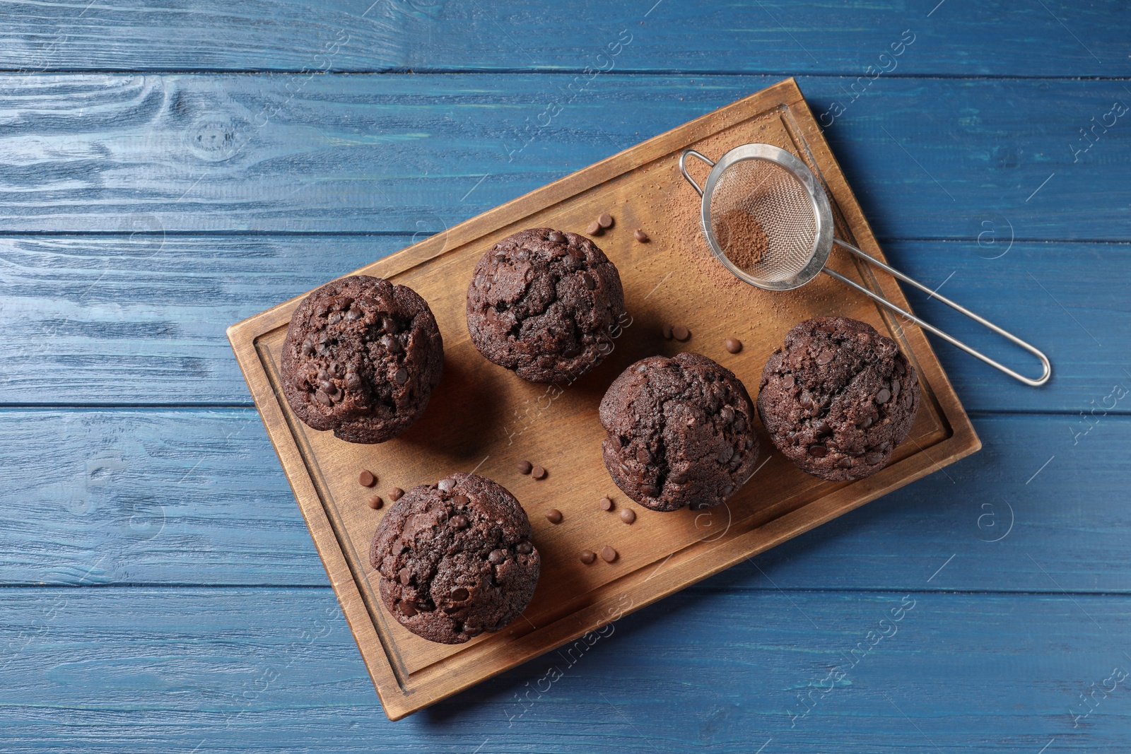 Photo of Delicious chocolate muffins and sieve with cocoa powder on blue wooden table, top view