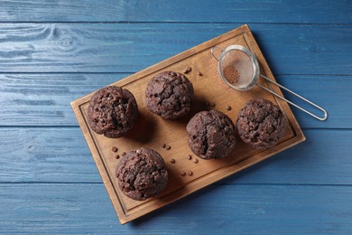 Delicious chocolate muffins and sieve with cocoa powder on blue wooden table, top view