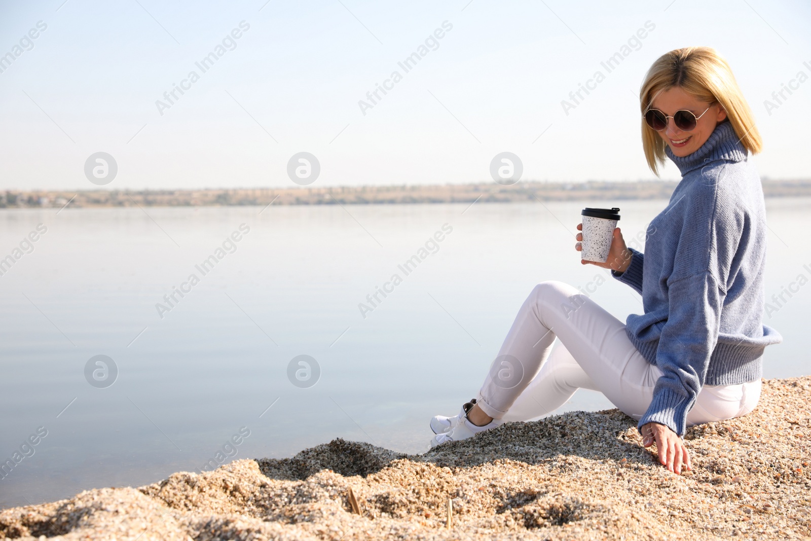 Photo of Happy woman in stylish sweater sitting on beach with cup of coffee