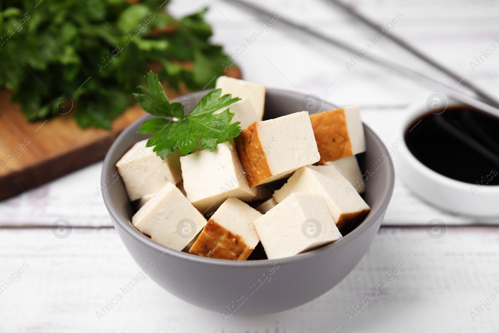 Photo of Bowl of smoked tofu cubes, soy sauce and parsley on white wooden table, closeup